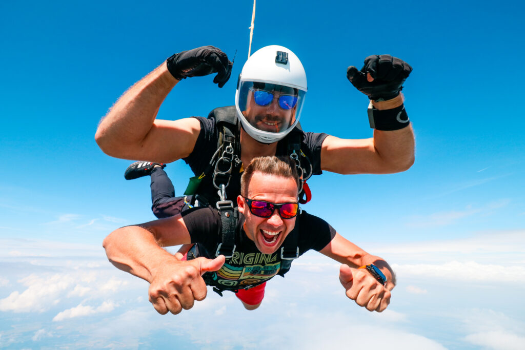 Tandem skydiving passenger with sunglasses giving a thumbs up in free fall