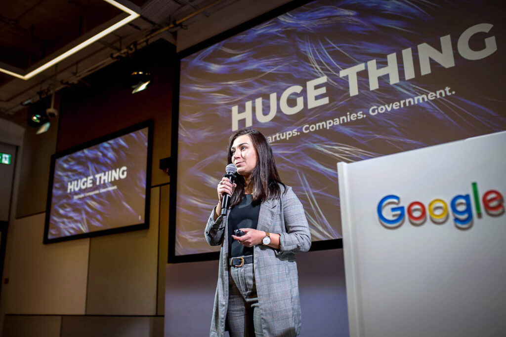 Speaker on stage at a Huge Thing event, with a Google sign beside her