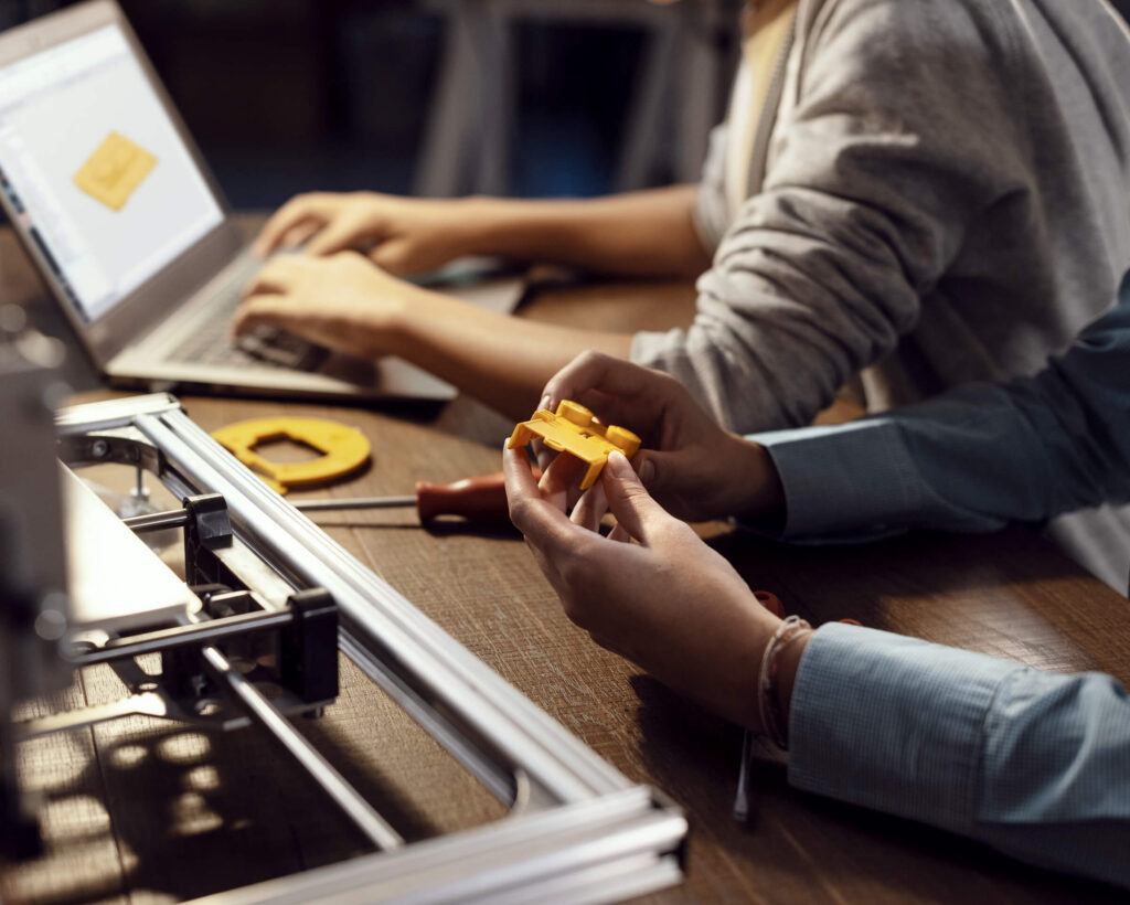 Hands holding a 3D printed part next to a 3D printer and a laptop
