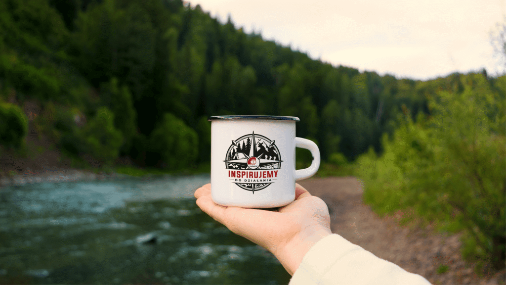 Enamel mug with the "Inspirujemy do działania" emblem held over a river