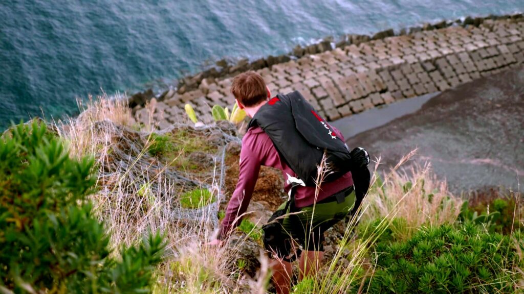 BASE jumper climbing through grass on a cliff above the sea, Madeira