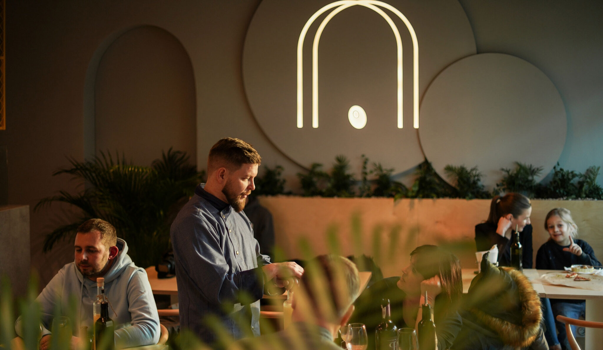 Guests and a waiter in the Ukryta Olivka pizzeria, with the arch logo as a neon sign