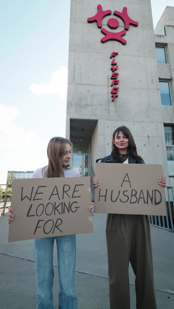 Flyspot reel still: two women holding signs "We are looking for a husband" outside the wind tunnel
